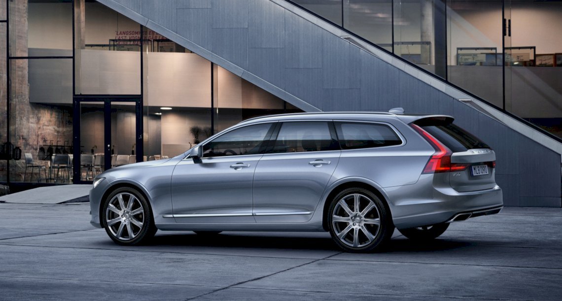 Volvo V90 INSCRIPTION in Bright Silver Metallic steht neben einem modernen, beleuchteten Haus auf einem Steinboden - Heckseitenschuss