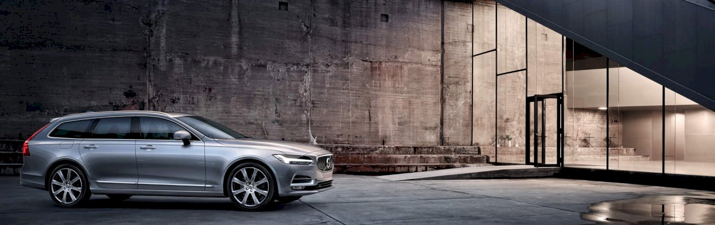 Volvo V90 INSCRIPTION in Bright Silver metallic steht auf einem Betonboden vor einer Steinwand, neben einem beleuchteten Raum - Frontseitenschuss